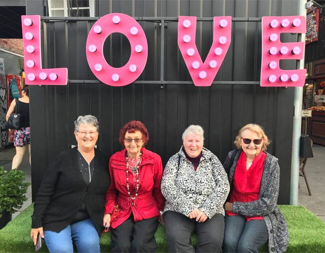 Four women sitting on a bench in front of a sign that says love during an over 50s tour in Australia.