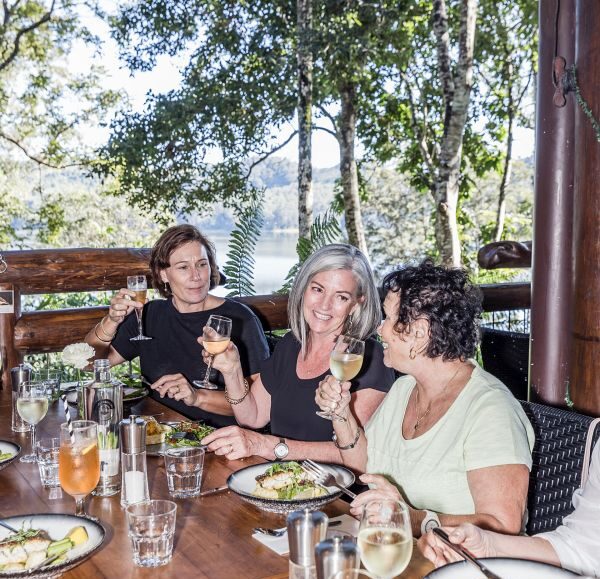 Three women enjoying a 3-course lunch outdoors, toasting with glasses of wine at a wooden table with plates of food, surrounded by lush greenery and a view of the lake at Secrets on the Lake.